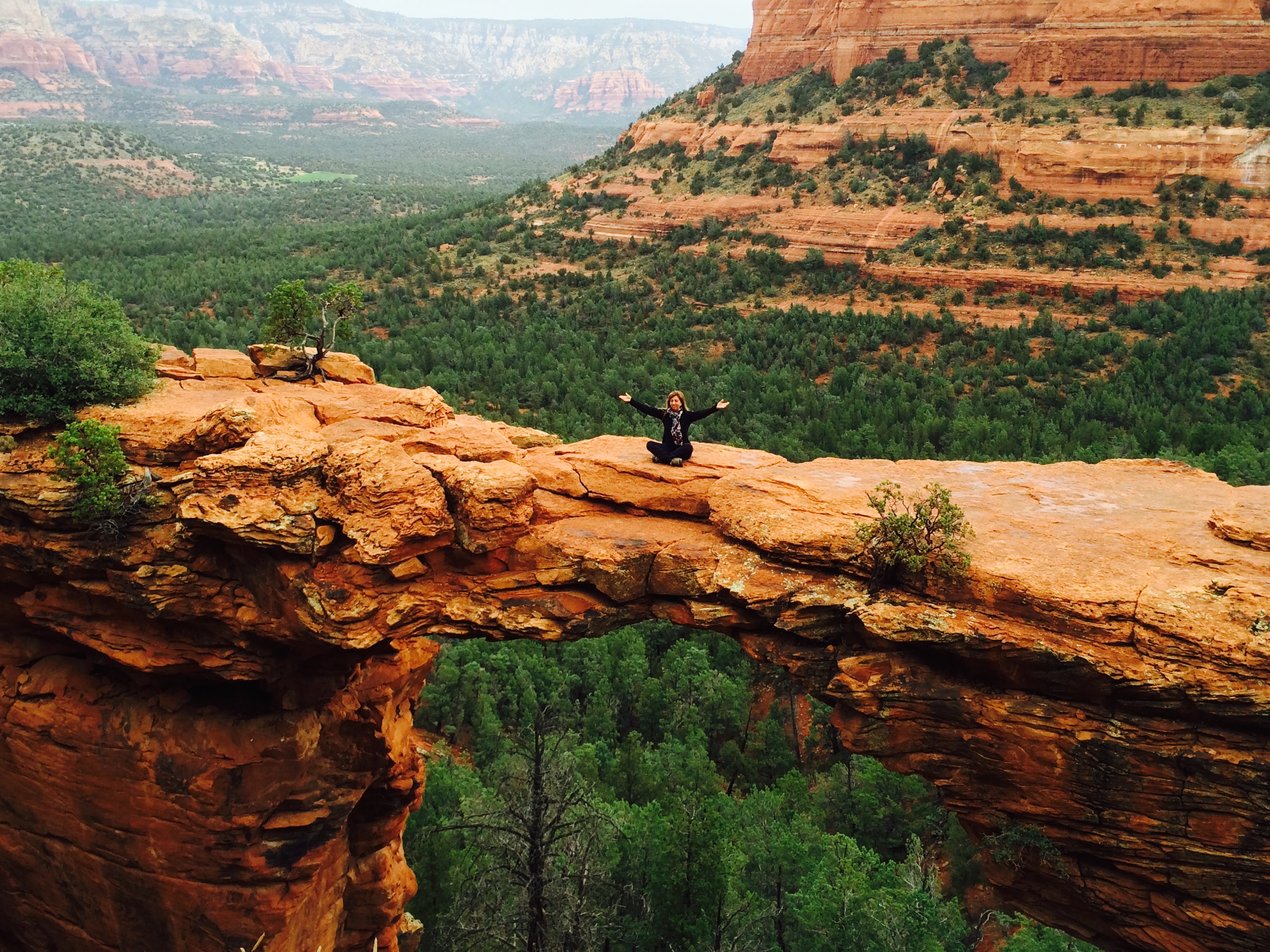 A guest seated with open arms on a natural sandstone arch overlooking the Sedona forest